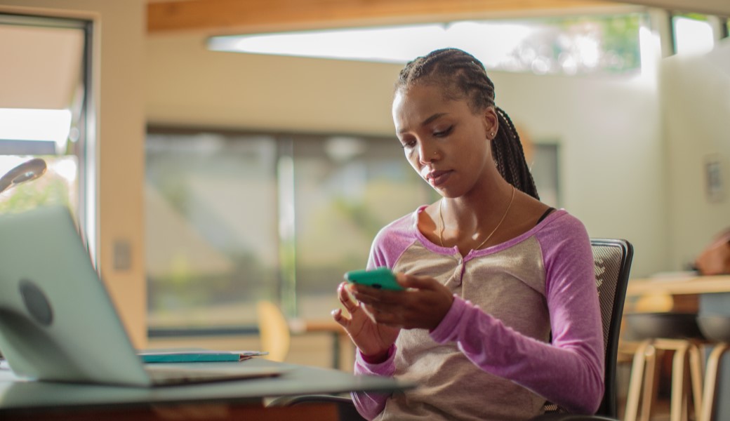 A woman scrolling on the phone while sitting on the chair in front of the laptop