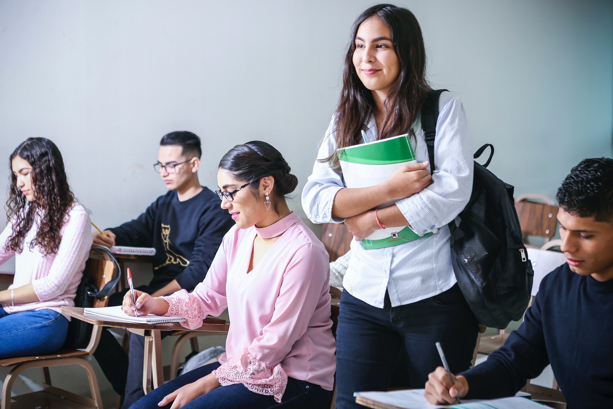 Student holding her books, standing and smiling in a classroom with peers.