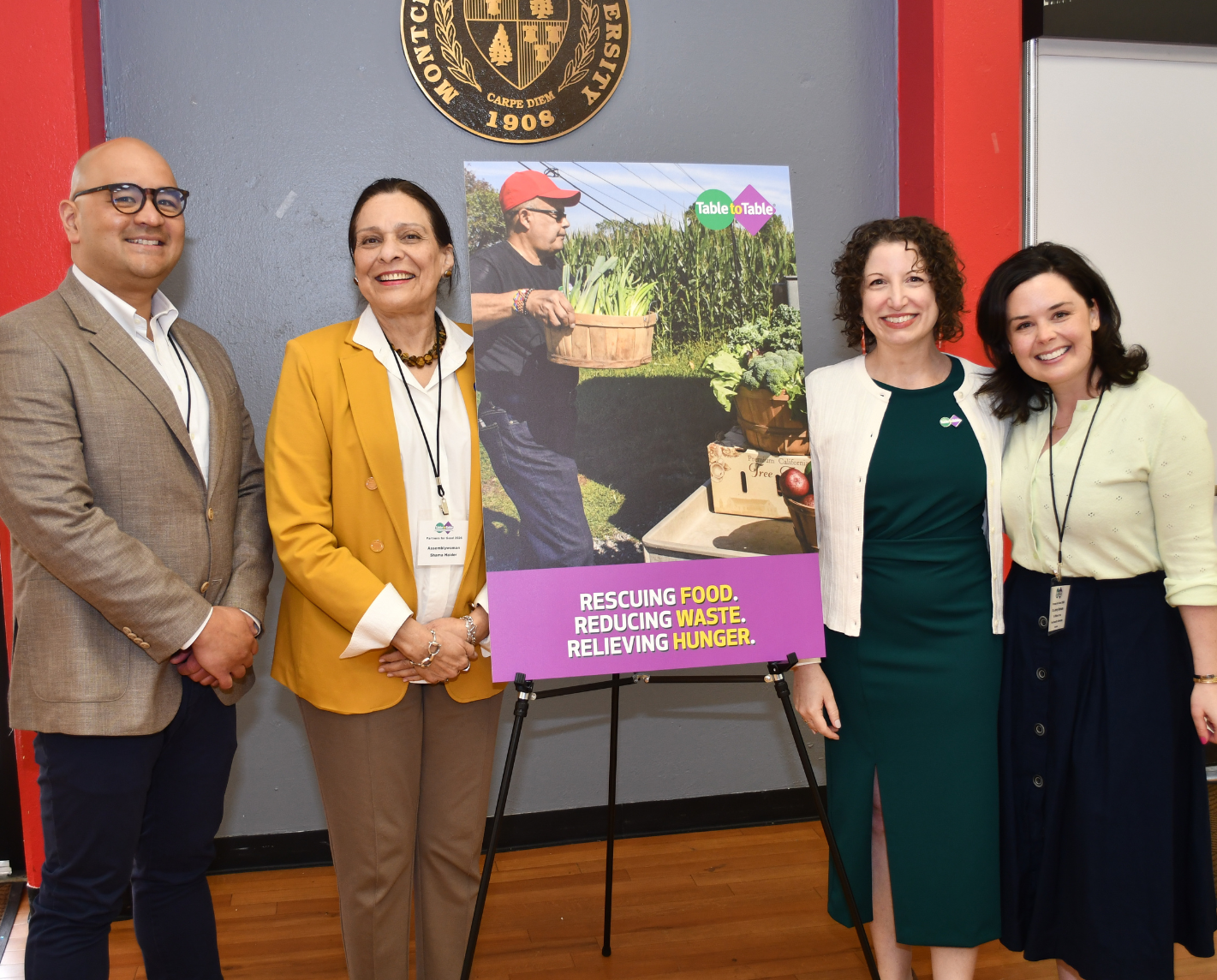 Mark Dinglasan, executive director of the New Jersey Office of the Food Security Advocate, Assemblywoman Shama Haider, Heather Thompson, executive director of Table to Table, and Dr. Jenny Schrum, of the New Jersey Office of the Food Security Advocate smile beside a colorful poster on an easel. The poster displays a farmer with harvested vegetables and text: "Rescuing Food. Reducing Waste. Relieving Hunger."