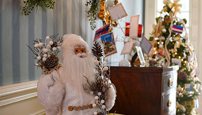 photo - The Foyer, by Keyport Garden Club, features numerous elegant decorations including a white-robed Santa Claus figure and a large wall spray of greenery with gold and cream colored balls and pinecones.