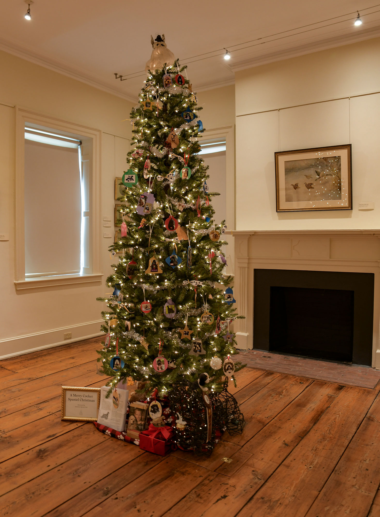 “A Merry Cocker Spaniel Christmas” tree by the American Spaniel Club/Southern NJ features hand made ornaments of Cocker Spaniels among bright lights. The room is beige with a wildlife painting above a fireplace.