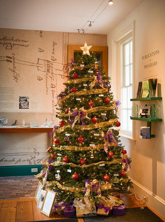photo - The “Spirit of the Present Day” Christmas tree stands at the rear entryway to Morven. A festive gold ribbon wraps around many colors of special and cheerful ornaments and bows. Museum displays can be seen  to the right and to the left.