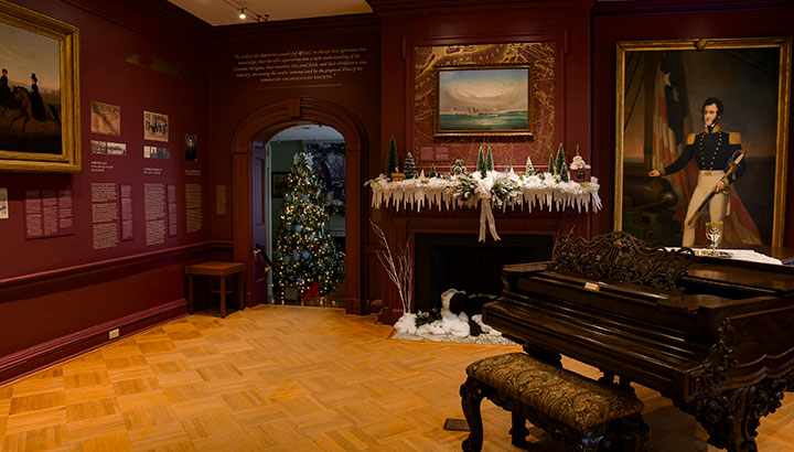 photo - A winter-themed mantelpiece by Neshanic Garden Club is shown in the music room. Plum colored walls, historic paintings and a grand piano are prominent. In the background entry to another room where a Christmas tree is lit.