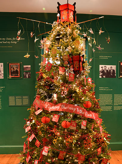 photo - Mount Laurel Garden Club tree called “Chinese New Year” is seen in a room with dark green walls featuring historical information. Bright red garlands wrap around with the center ribbon reading Lighting the Way to the New Year.