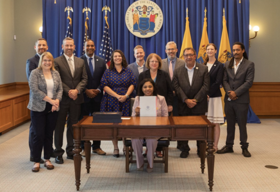 Acting Governor Tahesha Way showing the signed law A2929/S1034 with a team on the background