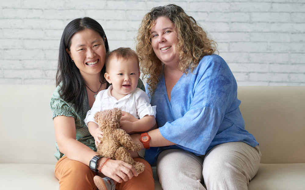 A mixed-race same-sex female couple sitting on a sofa holding their smiling baby