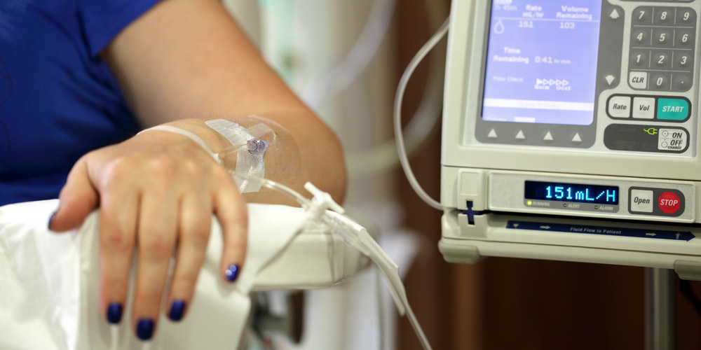 A woman's hand resting on a chair in a hospital room, with an IV going into it