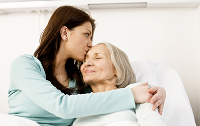 A smiling elderly woman in a hospital bed being hugged and kissed by her daughter