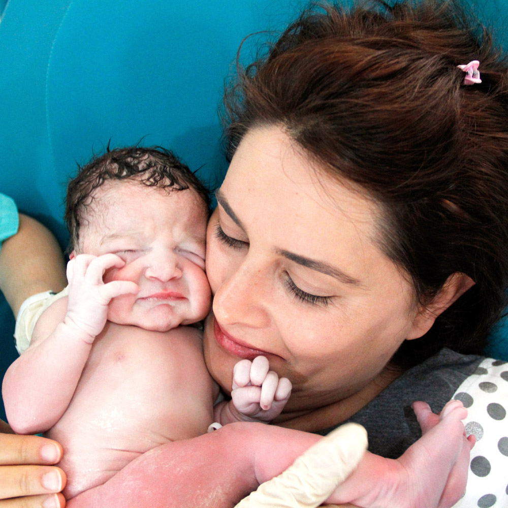 A woman in a hospital bed who just gave birth smiling at the baby, who is being held close to her face by a doctor