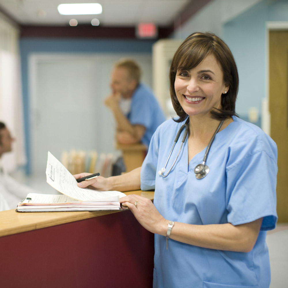 A female doctor smiling and leaning against a nurse's station desk at a hospital