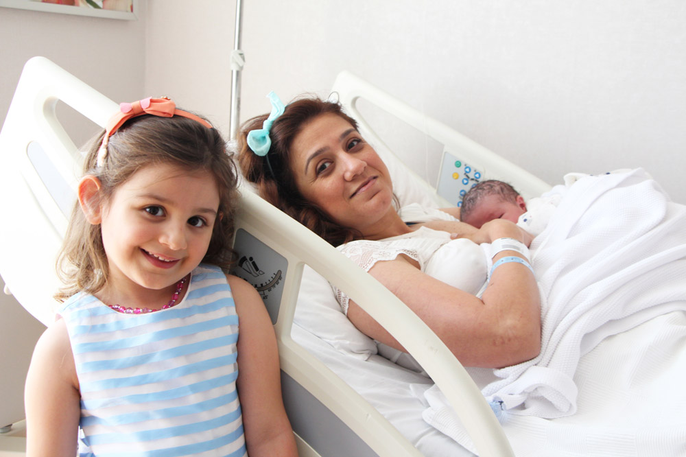 Mother in hospital bed holding newborn, with an older child standing next to the bed, smiling