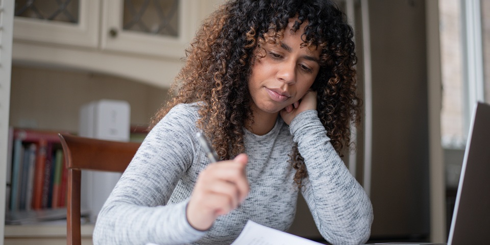 A young woman sits at her kitchen counter alone and fills out forms with a pen. Her laptop is open on the counter.