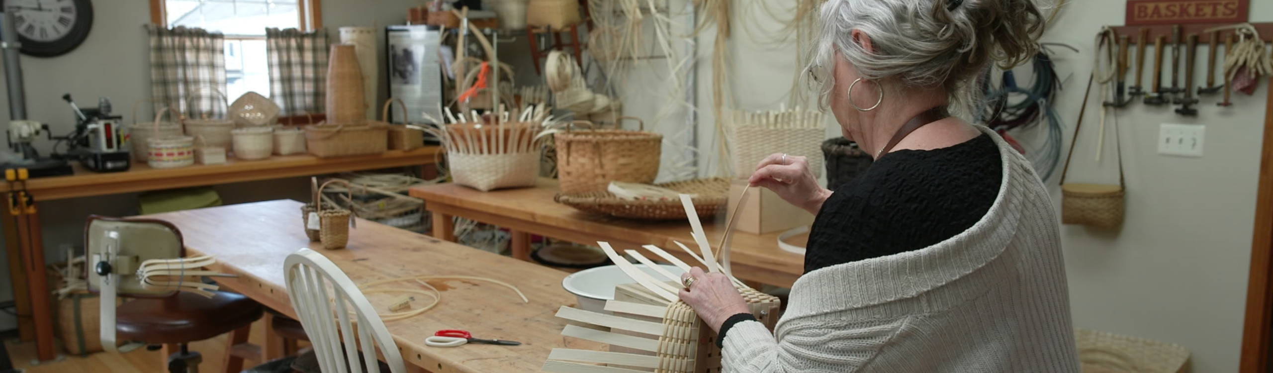 a woman with silver hair weaves a traditional basket in her studio