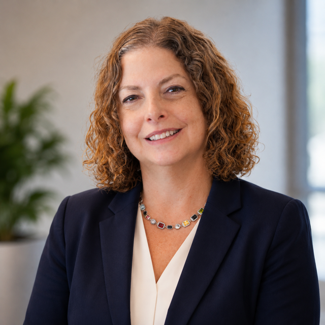 A headshot of a woman with shoulder-length curly brown hair, smiling warmly. She is wearing a dark blue suit jacket over a white V-neck blouse. She is wearing a silver necklace with different color stones all around it including red, blue, and yellow stones. The background of the image is blurred, a room with a gray wall can be seen behind her. To the left of the woman is a large green plant in a white pot. To her right is a window with sunlight coming through.