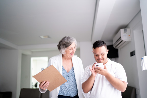 Adult man with an intellectual disability standing next to his female doctor in a medical office with white walls. The man has short brown hair and is wearing a white shirt. He is looking at his cell phone. The female doctor is looking at the man’s cell phone. She has short curly white hair and is wearing dark pants, a light blue shirt, and a white lab coat. She is holding a clipboard.